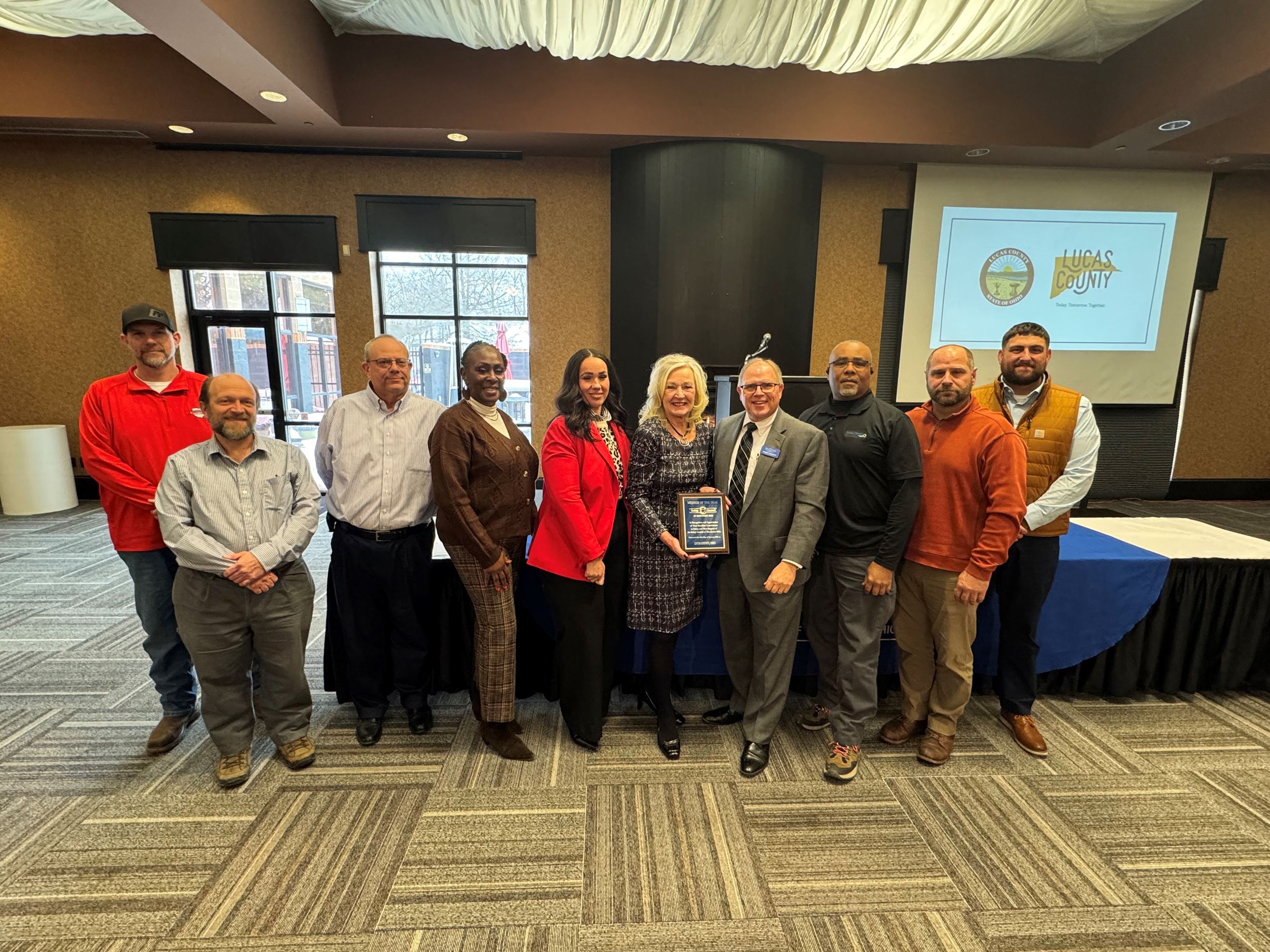 Ten people stand in a row in a conference room holding an award.